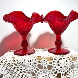May include: Two matching red glass compote dishes featuring a diamond-patterned bowl with a ruffled, scalloped rim. Each piece stands on a pedestal base with a textured stem and a circular foot, showcasing a vibrant, translucent ruby-red finish.