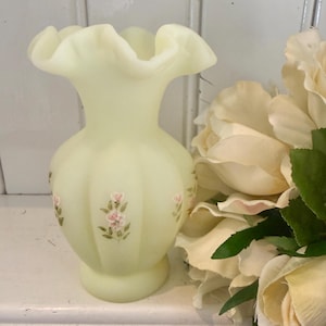 May include: A pale yellow satin glass vase with a ruffled, flared rim and a bulbous, ribbed body. The exterior features hand-painted floral motifs consisting of small pink blossoms and green leaves arranged in vertical patterns around the centre.
