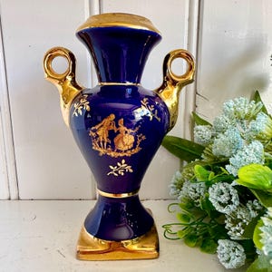 May include: A cobalt blue ceramic vase featuring gold-toned handles and a gold-toned base. The front of the vase displays a gold-coloured illustration of two figures in period clothing, surrounded by delicate gold leaf and vine patterns.