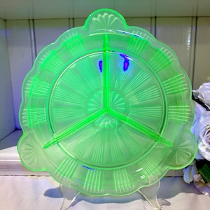 May include: A vintage, three-sectioned, green glass serving dish. The dish has a scalloped edge and a central design radiating outwards. The dish is supported by a clear stand. The glass has a slight glow.