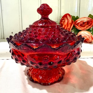 May include: A vintage ruby red glass candy dish with a lid. The dish has a hobnail pattern and a scalloped edge. The lid features a decorative finial. The dish sits on a pedestal base. Artificial roses are in the background.
