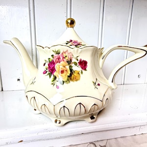 May include: A white porcelain teapot with a gold trim and floral design. The teapot has a lid with a gold knob and a spout on the side. The teapot is decorated with pink, yellow, and red roses.