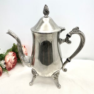 May include: A silver teapot with an ornate design and three legs. The teapot has a lid with a finial and a handle with a floral design.