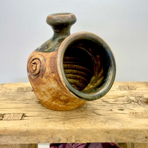 May include: A handcrafted ceramic vase with a unique, organic shape. The vase features a brown and tan colour palette with a dark green rim and accents. A spiral design is etched on the side. The vase is a decorative piece for the home.