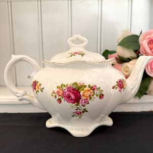 May include: A white porcelain teapot featuring a raised embossed scroll pattern and a colourful floral motif of pink, red, and yellow roses. The teapot includes a matching lid with a decorative handle and a scalloped base, finished with gold trim.