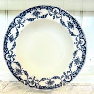 May include: White ceramic bowl with a scalloped edge and a blue floral pattern around the rim. The bowl has a wide, shallow shape and is displayed on a clear stand. The blue design includes floral and scroll motifs.