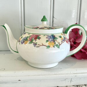 May include: A white ceramic teapot featuring a floral pattern with pink, blue, and yellow flowers on the body and lid. The teapot has a green rim on the lid, spout, and handle, and a small green finial on top of the lid, with a rounded base.