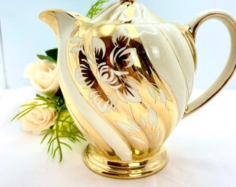 May include: A white teapot with gold accents and a floral design. The teapot has a white lid with a gold knob and a gold handle.