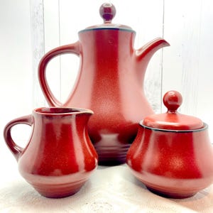 May include: A red ceramic tea set comprising a teapot, a creamer jug, and a lidded sugar bowl. The teapot has a curved handle and spout. Each piece features a dark rim and a rounded form. The set is displayed on a white surface.