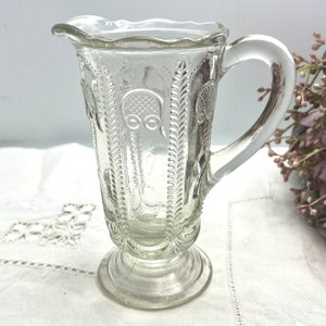 May include: A clear glass jug with a decorative embossed pattern. The jug has a rounded body, a flared base, and a handle.
