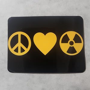 May include: Black rectangular item with yellow symbols: a peace sign, a heart, and a radiation symbol. The symbols are centered and evenly spaced on the black background.