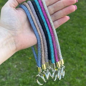 Può includere: Un set di cinque portachiavi da polso dal design tubolare ricoperto di piccoli strass scintillanti. I cinturini sono disponibili nei colori blu, champagne, nero, turchese e magenta, ciascuno rifinito con una chiusura e un anello in metallo color argento.