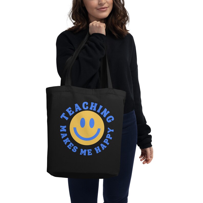 Pu&ograve; includere: Borsa tote nera con un'emoji sorridente gialla e la scritta blu "TEACHING MAKES ME HAPPY". La borsa &egrave; tenuta da una persona.