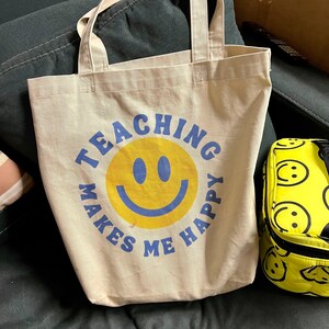 Pu&ograve; includere: Borsa tote beige con la scritta "TEACHING MAKES ME HAPPY" in blu attorno a una faccina sorridente gialla. Un portapranzo giallo con un motivo a faccina sorridente &egrave; visibile a destra.