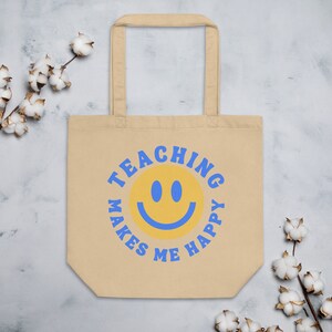 Pu&ograve; includere: Borsa tote beige con testo blu e una faccina sorridente gialla. Il testo recita "TEACHING MAKES ME HAPPY" in un motivo circolare attorno alla faccina sorridente. La borsa ha due manici ed &egrave; realizzata in un materiale simile alla tela.