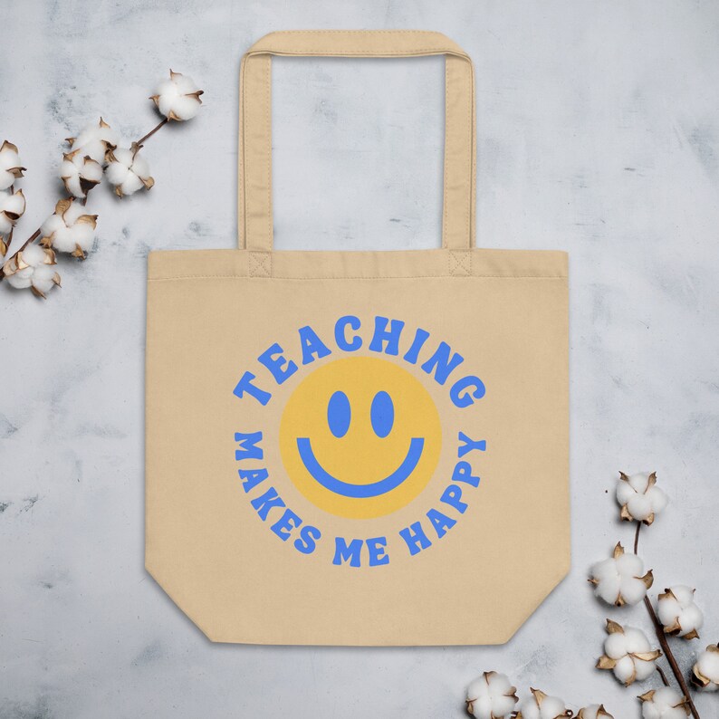 Pu&ograve; includere: Borsa tote beige con testo blu e una faccina sorridente gialla. Il testo recita "TEACHING MAKES ME HAPPY" in un motivo circolare attorno alla faccina sorridente. La borsa ha due manici ed &egrave; realizzata in un materiale simile alla tela.