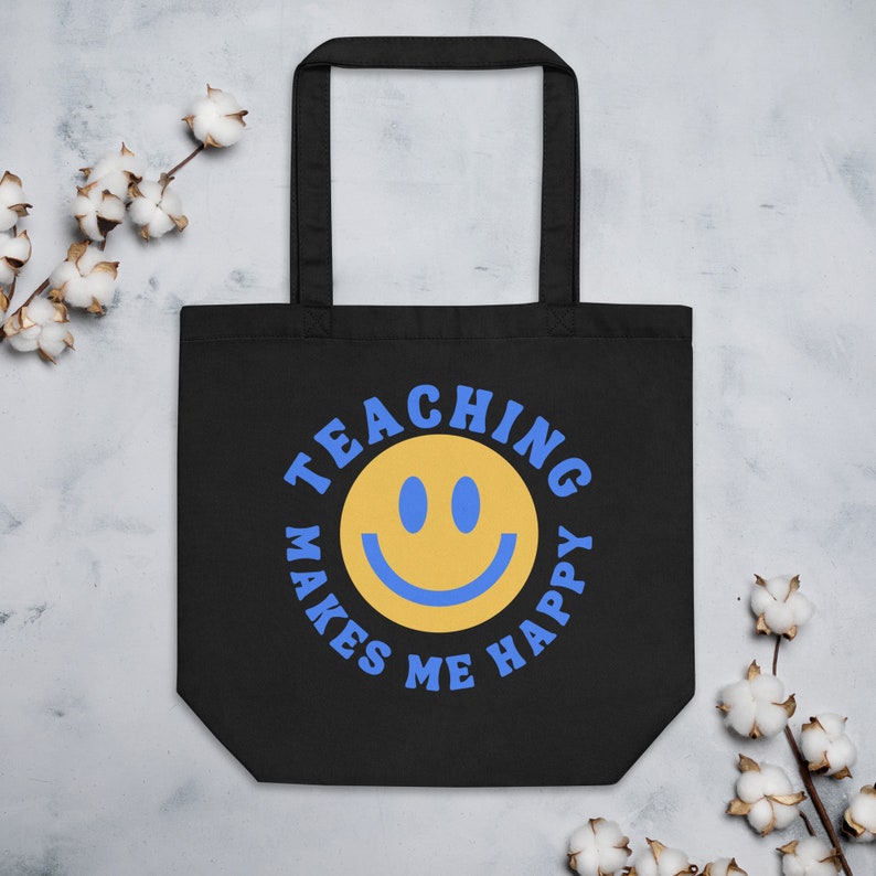 Pu&ograve; includere: Borsa tote nera con un'emoticona gialla e la scritta blu "TEACHING MAKES ME HAPPY". La borsa ha due manici neri ed &egrave; realizzata in materiale resistente. Il design &egrave; centrato sulla parte anteriore.