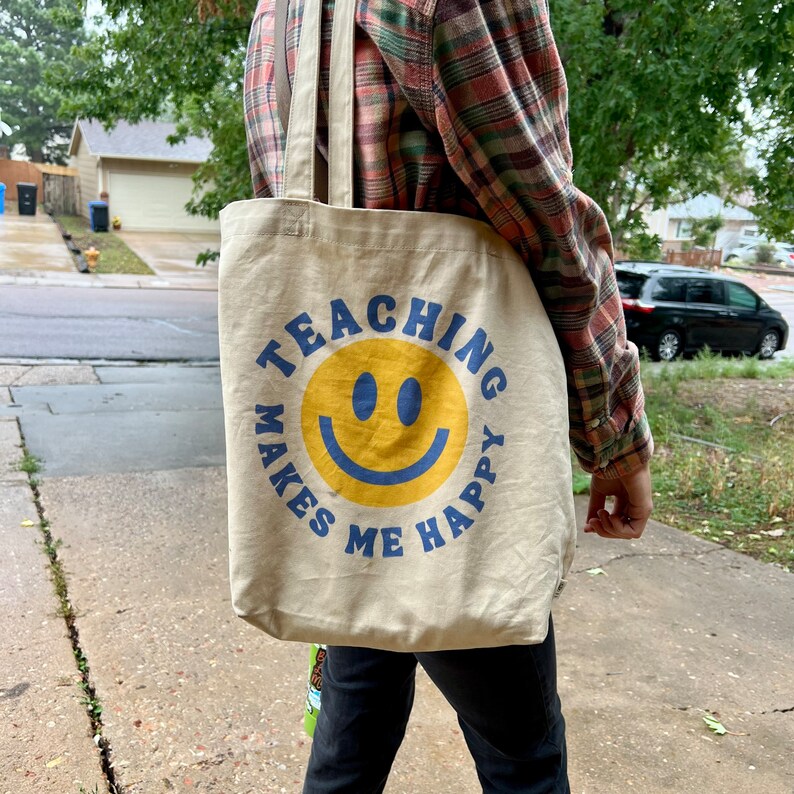 Pu&ograve; includere: Borsa tote in tela beige con la scritta "TEACHING MAKES ME HAPPY" in blu, che circonda una faccina sorridente gialla. La borsa ha lunghe cinghie e viene trasportata all'aperto.