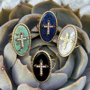 Peut inclure: Quatre bagues ovales avec des montures en métal doré aux bords perlés. Chaque bague présente une incrustation de croix centrale ornée de petites pierres claires. Les pierres sont placées sur quatre fonds distincts : vert clair, bleu, blanc et noir.