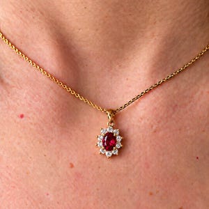 Peut inclure: Un collier à chaîne dorée avec un pendentif orné d'une pierre rouge centrale de forme ovale, entourée d'un halo de douze petites pierres claires à taille ronde. Le pendentif est suspendu à une fine chaîne à maillons dorés sur la peau.
