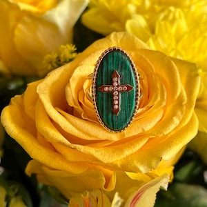Puede incluir: Un anillo ovalado con una piedra de malaquita verde y una incrustación de cruz en tono dorado con pequeñas piedras claras. El anillo tiene un borde de metal dorado con cuentas y una banda fina, centrado sobre una flor de rosa amarilla.
