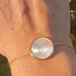 Peut inclure: Bracelet doré avec un grand pendentif rond en nacre irisée. Le pendentif est serti dans une lunette dorée et relié à une chaîne délicate. Le bracelet est porté sur un poignet, captant la lumière du soleil.