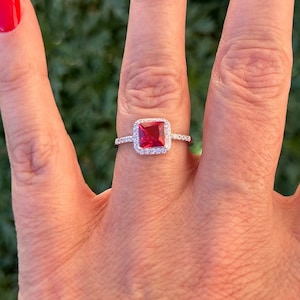Può includere: Un anello d'argento con una pietra rossa quadrata circondata da un alone di piccole pietre bianche.