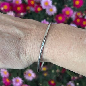 Puede incluir: Un brazalete delgado de color plateado. El metal liso y curvado refleja la luz, creando una estética brillante y minimalista. El brazalete se muestra sobre un fondo de vibrantes flores rosas y amarillas.