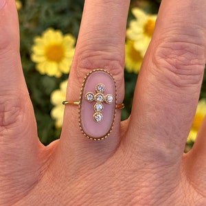 Puede incluir: Un anillo de tono dorado con una piedra cabujón rosa de forma ovalada engastada en un borde de metal con cuentas. El centro de la piedra rosa está adornado con un pequeño motivo de cruz compuesto por cinco piedras redondas transparentes.