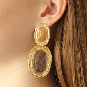May include: Gold-toned, two-part earrings with oval-shaped, textured frames. The top oval features a light brown, marbled design, while the bottom oval has a darker brown, swirling pattern. The earrings are a statement piece.