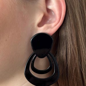 Peut inclure: Une boucle d'oreille noire brillante composée d'une pièce supérieure solide en forme de goutte d'eau reliée à deux anneaux concentriques en forme de goutte d'eau avec un centre ouvert. La surface est lisse, réfléchissante et géométrique.