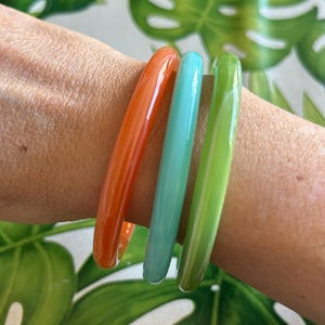 Op de afbeelding: Drie kleurrijke armbanden in oranje, turquoise en groen. De gladde, glanzende armbanden zijn op een pols gestapeld. De achtergrond heeft groene en witte tropische bladeren. Deze armbanden zijn een levendig accessoire.