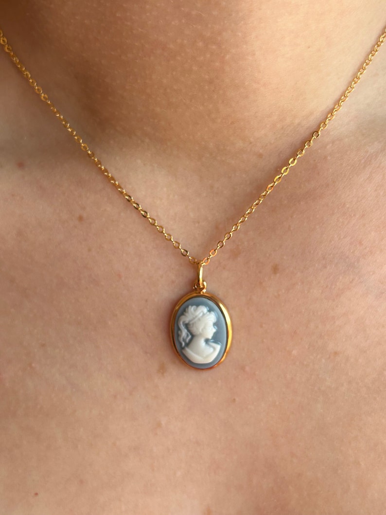Pu&ograve; includere: Una collana color oro con un ciondolo a cammeo raffigurante il profilo di una donna. Il cammeo &egrave; incastonato in una cornice ovale con sfondo azzurro chiaro e profilo bianco. La collana &egrave; esposta su uno sfondo color pelle.