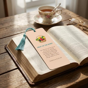 Op de afbeelding: Een perzikkleurige bladwijzer met een turquoise kwast ligt op een open boek. De bladwijzer heeft een illustratie van een fruitmand en de tekst "FRUIT OF THE SPIRIT" en een bijbelvers. Een theekopje en een bril staan op de achtergrond.