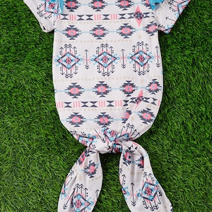 May include: A white baby gown with a pink, blue, and gray geometric pattern. The gown has short sleeves with blue ruffles and a tie at the bottom.