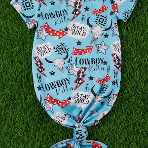 May include: A light blue baby gown with a white and red pattern. The pattern features the words "Cowboy Killer" and "Stay Wild" repeated throughout. The gown has a knotted tie at the bottom.