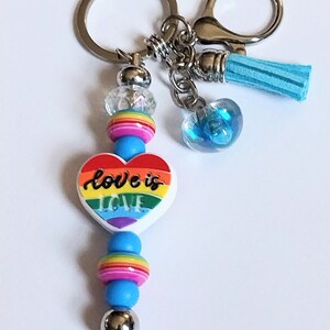 May include: A keychain with a silver ring and clasp, featuring a heart-shaped charm with a rainbow design and the words "love is love." The keychain also includes blue beads, a clear heart-shaped charm, and a blue tassel.