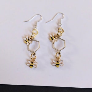 May include: A pair of silver and gold earrings featuring a hexagonal honeycomb design with two small bee charms dangling from each earring.