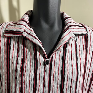 May include: A collared shirt with vertical stripes in grey, black, and red. The fabric has a textured, pleated appearance. The shirt has a button closure at the neckline.