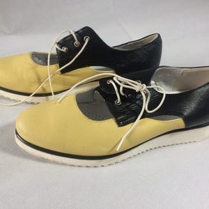 Może przedstawiać: Para dwukolorowych butów damskich. Buty mają żółtą skórzaną cholewkę i czarną skórzaną piętę. Posiadają białe sznurowadła i białą podeszwę z czarnym wykończeniem.