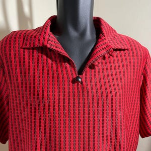 May include: Red short-sleeved shirt with a black vertical pattern. The shirt has a collared neckline and a single black button closure. The fabric has a textured appearance, and the shirt is displayed on a black mannequin.