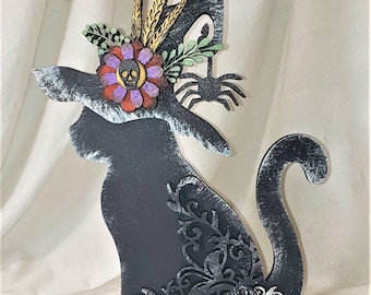 May include: Black wooden cat figurine with a witch's hat adorned with a skull and floral accents. The cat is sitting on a black base.