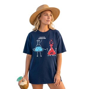 May include: A navy blue t-shirt featuring a graphic of two laboratory flasks with faces. One blue flask says, I THINK YOU'RE OVERREACTING, while the red flask next to it is bubbling over. The shirt has short sleeves and a crew neckline.
