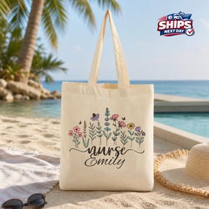 May include: A beige tote bag with a floral design and the text "nurse Emily". The flowers are in shades of pink, purple, yellow, and blue. The bag has two handles and is sitting on a sandy beach.