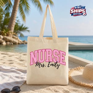 May include: A beige canvas tote bag with the word "NURSE" in pink block letters and the name "Mrs. Emily" in black script. The bag has two handles and is sitting on a sandy beach with sunglasses and a sun hat.