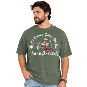 May include: An olive green short-sleeve t-shirt featuring a graphic of a red steam locomotive surrounded by pine trees. The text reads All Aboard Admit One Dec. 25 Polar Express in white lettering across the chest area of the garment.