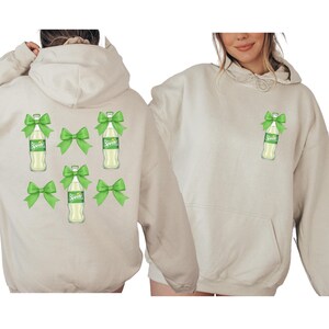 May include: A beige hoodie featuring a graphic of a Sprite bottle with a green bow. The back of the hoodie has multiple Sprite bottles with green bows. The front of the hoodie has a single Sprite bottle with a green bow.