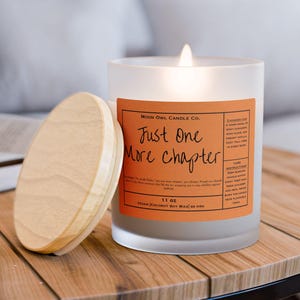Puede incluir: Una vela de vidrio esmerilado encendida con una tapa de madera está sobre una mesa de madera. La vela tiene una etiqueta naranja con el texto "Just One More Chapter". La vela es de 312 gramos.