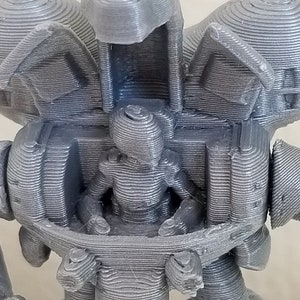 May include: A 3D printed model of a robot with a human figure sitting inside. The robot is gray and has a detailed design. The human figure is also gray and has a simple design.