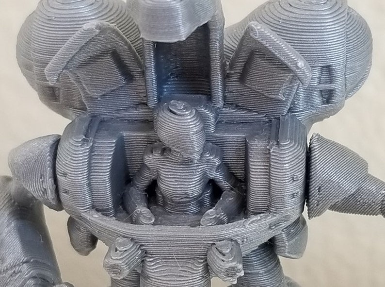 May include: A 3D printed model of a robot with a human figure sitting inside. The robot is gray and has a detailed design. The human figure is also gray and has a simple design.